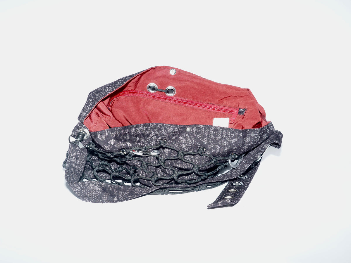 Handmade bag made of upcycled silk kimono and encased in a hand-knotted black paracord. The fabric features purply-black fabric with white stitching and a red interior. The strap includes grommets and a carabiner for adjustability.